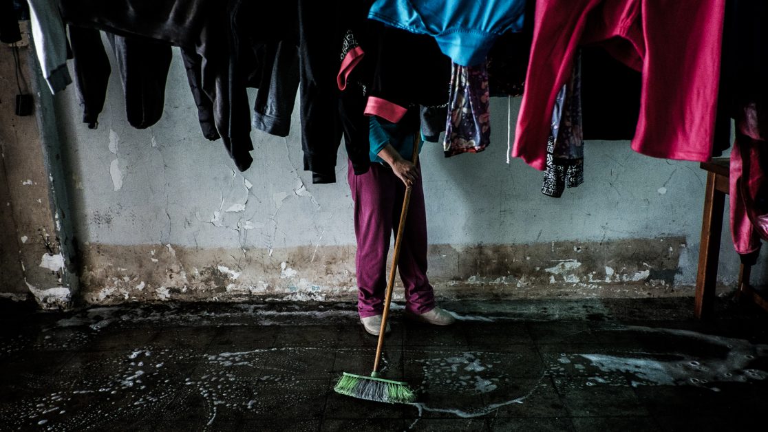 Una mujer escobilla el suelo entremedio de la ropa colgada en el primer piso. Foto: Jorge Vargas | Migrar Photo